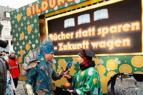 Interaktiver Mottowagen „Bildungs-Casino“ der Hochschule Düsseldorf beim Rosenmontagszug: Eine Person im Meereskostüm interviewt Professorin Felicitas Rohden vor einer großen LED-Wand mit der Aufschrift „Bücher statt sparen – Zukunft wagen“.